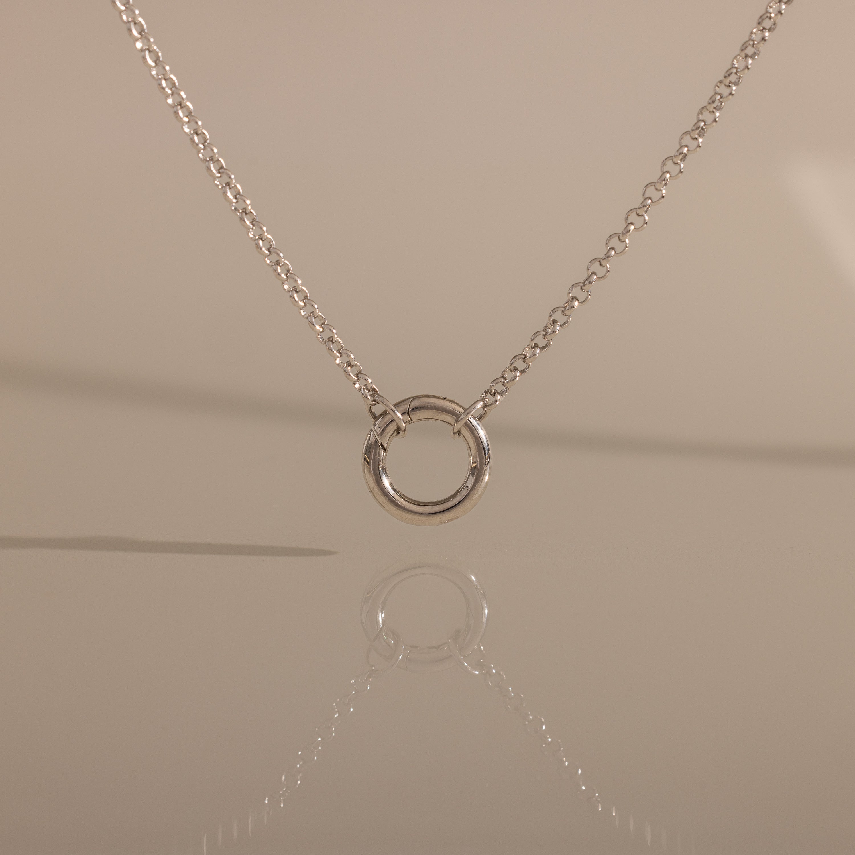 The Cable Circle Charm Necklace in Silver features a simple silver chain with a small circular pendant, elegantly reflected on a smooth surface.