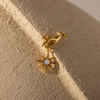 The Star Opal Hand Charm earring features a hand holding a star and opal stones, symbolizing intuition and cosmic energy, shown on a textured beige surface.