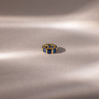 The Infinity Princess Sapphire Charm, a gold ring with princess-cut blue gemstones, is displayed on soft, light fabric, evoking elegance amid gentle shadows.
