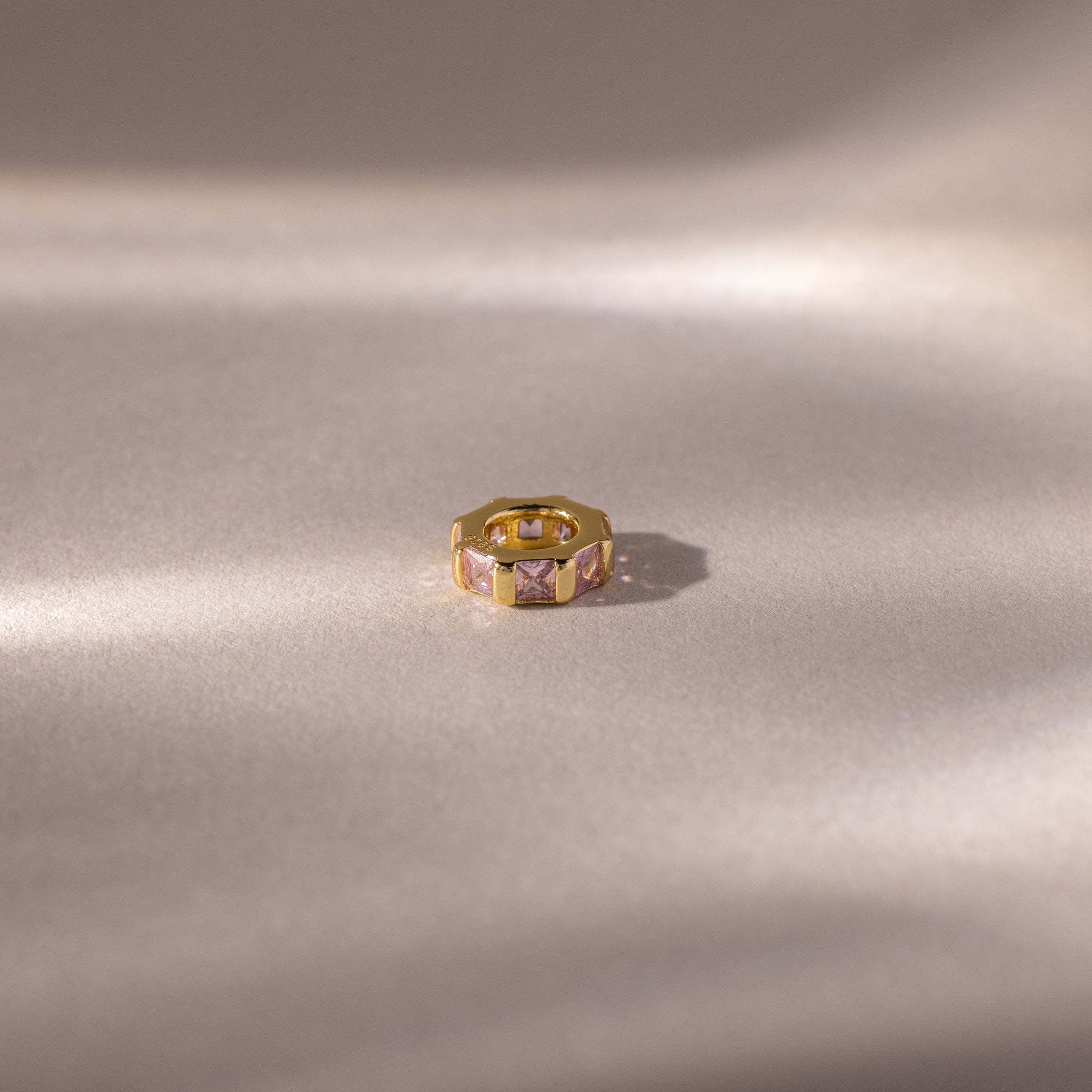 The Infinity Princess Pink Tourmaline Charm, featuring pink gem insets and delicate princess-cut stones, rests on a soft, light beige satin surface with gentle shadows.