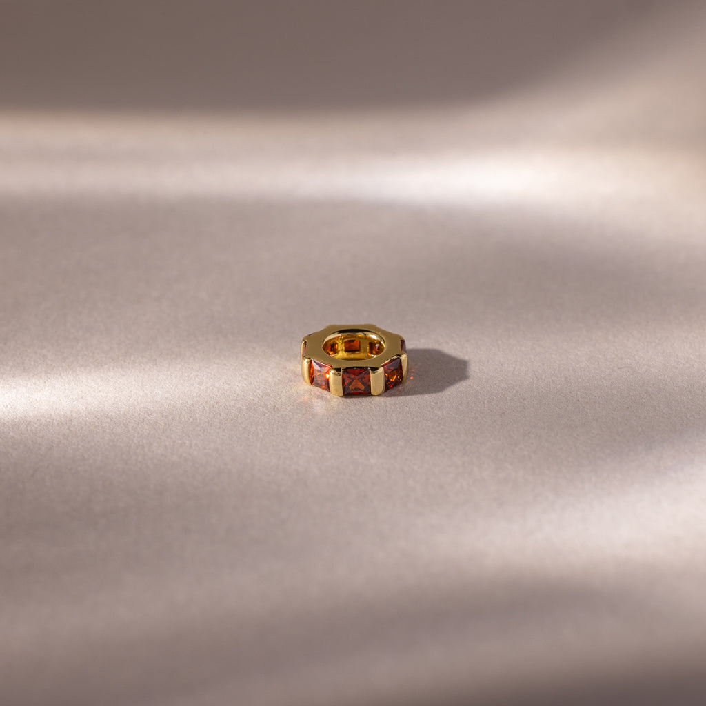 The Infinity Princess Garnet Charm, adorned with princess-cut stones and red garnet accents, rests on a softly lit, light-colored surface.