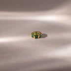 The Infinity Princess Emerald Charm, a gold ring with green princess-cut stones, rests on a soft, light beige surface with gentle shadows.
