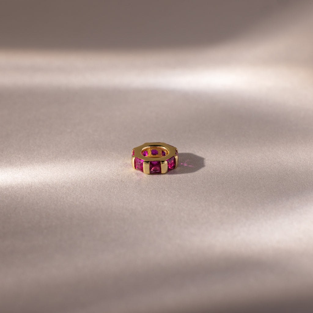 The Infinity Princess Ruby Charm is showcased on a soft, neutral surface with gentle lighting.