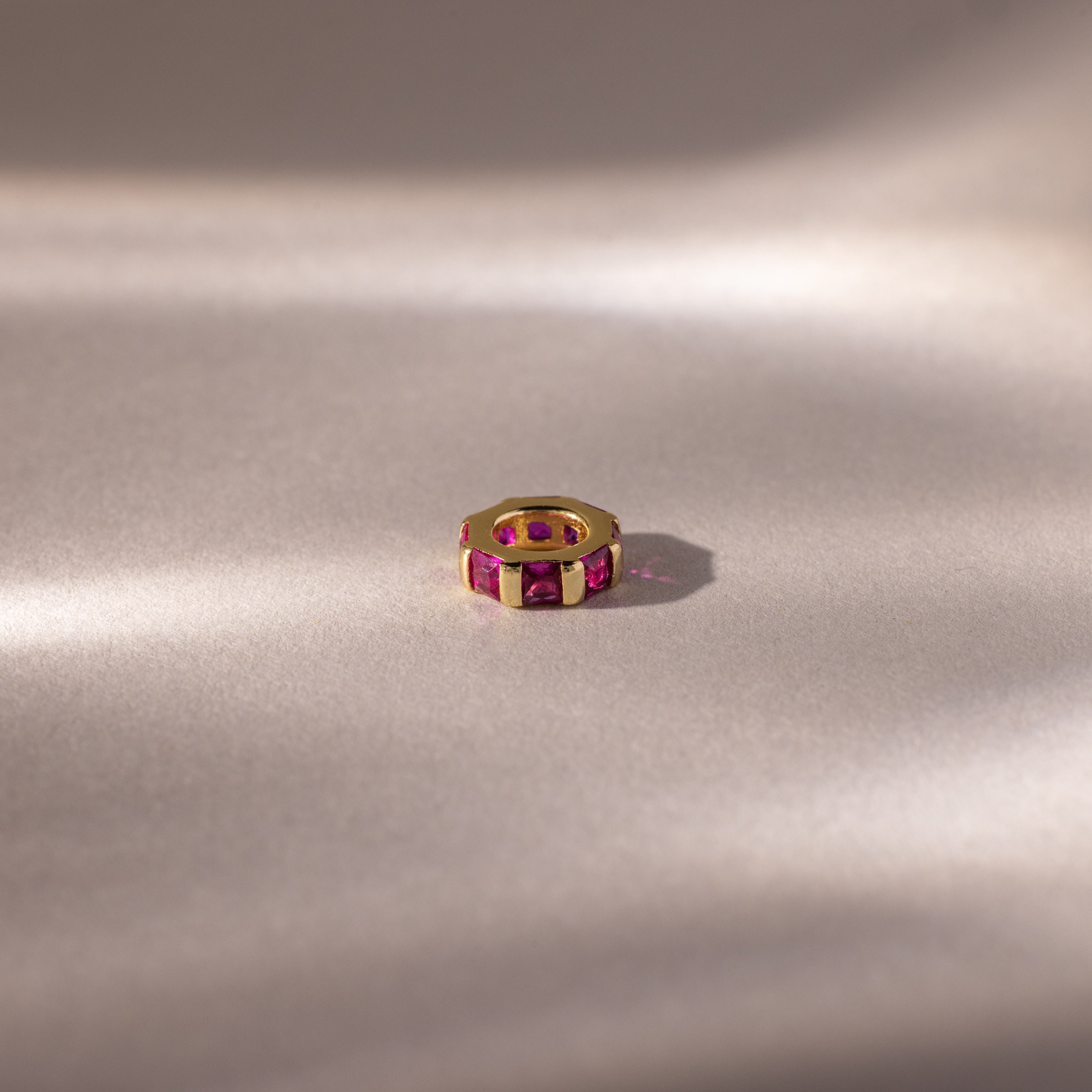 The Infinity Princess Ruby Charm is showcased on a soft, neutral surface with gentle lighting.