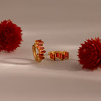 Staggered Baguette Garnet Huggies, featuring rectangular garnet stones, are shown between two red flowers on a neutral background, radiating modern elegance.