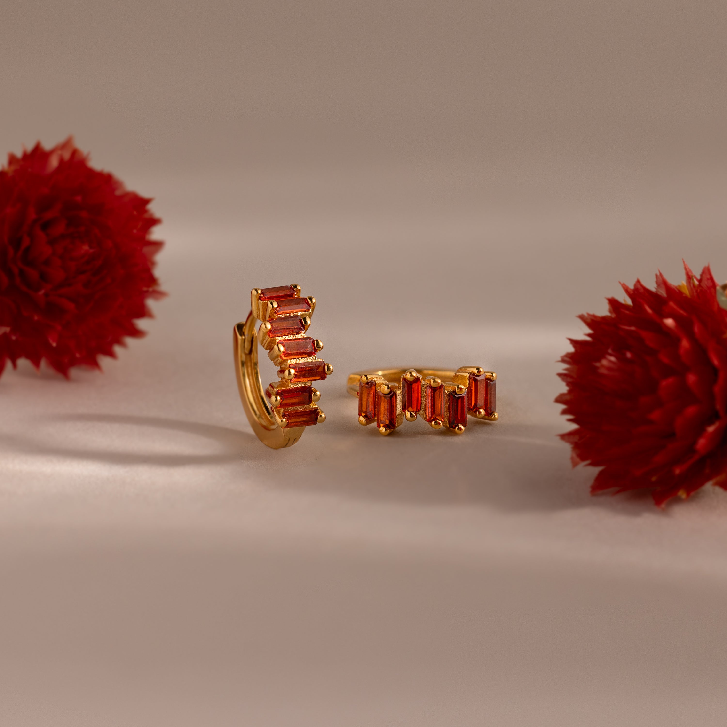 Staggered Baguette Garnet Huggies, featuring rectangular garnet stones, are shown between two red flowers on a neutral background, radiating modern elegance.