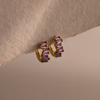 The Staggered Baguette Amethyst Huggies are modern gold hoops adorned with three rectangular amethyst stones on each, elegantly displayed on a textured beige surface.