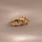 The Staggered Baguette Diamond Huggies, featuring gold hoops set with baguette-cut diamonds, rest on a soft beige surface—elegant modern earrings that add refined sparkle to any look.