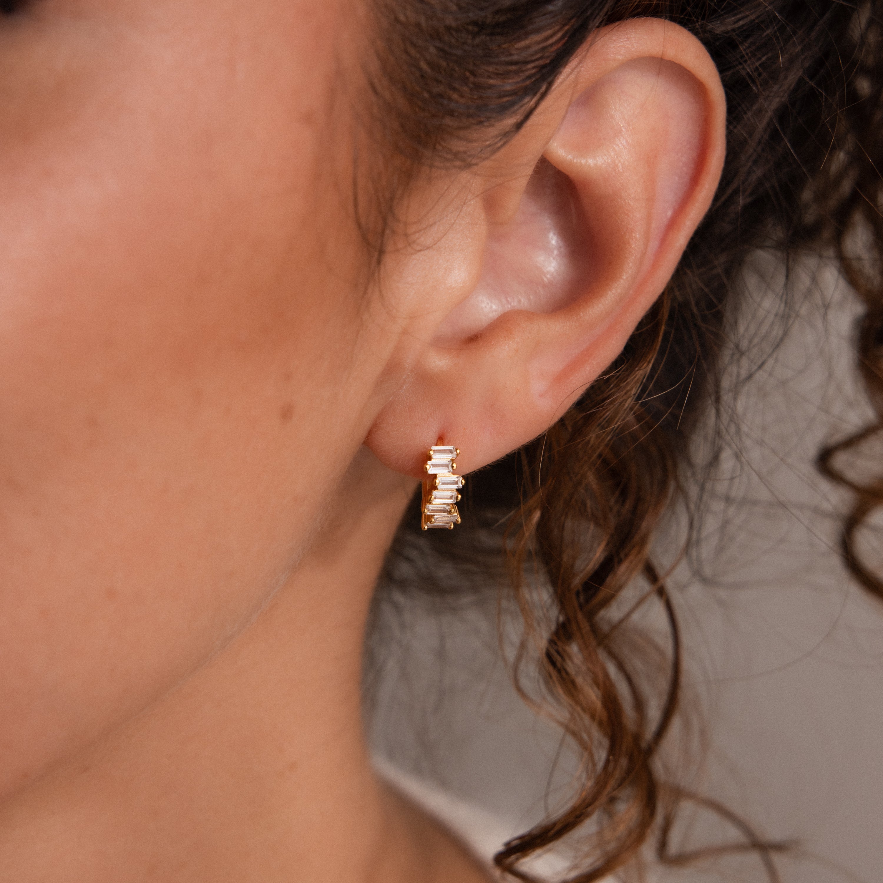 Close-up of a woman's ear wearing the Staggered Baguette Amethyst Huggies: small gold hoops with staggered baguette-cut amethyst stones for a chic, contemporary look.