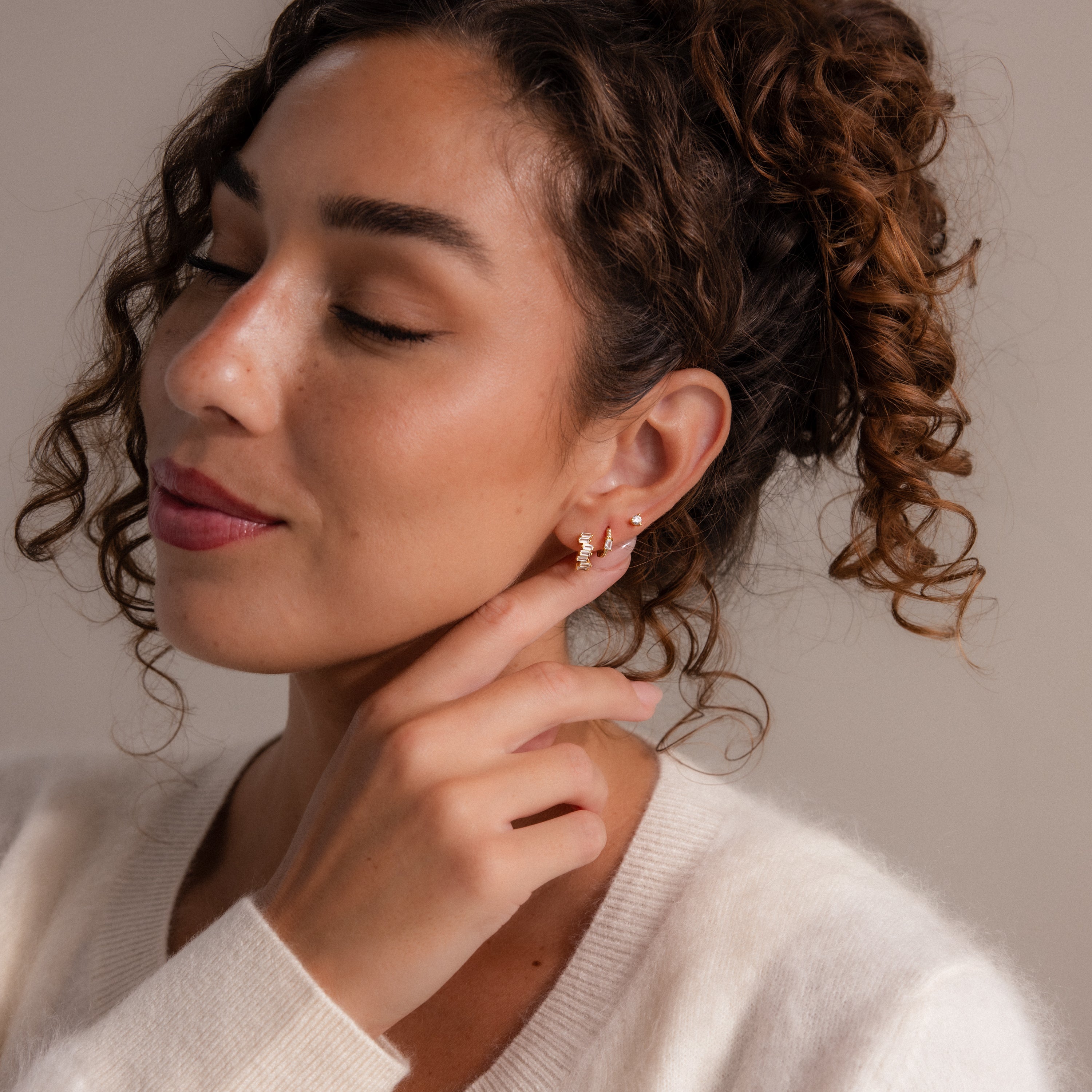 A woman with curly hair smiles softly, touching her gold Staggered Baguette Garnet Huggies, radiating modern elegance in a cream sweater.