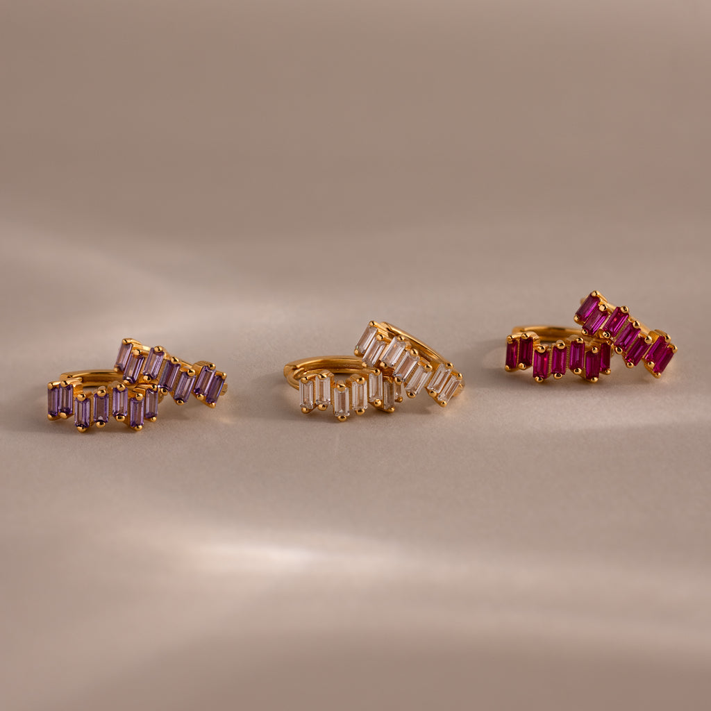 Three pairs of Staggered Baguette Birthstone Huggies, featuring purple, clear, and pink baguette-cut stones, showcase modern elegance on a soft beige background.