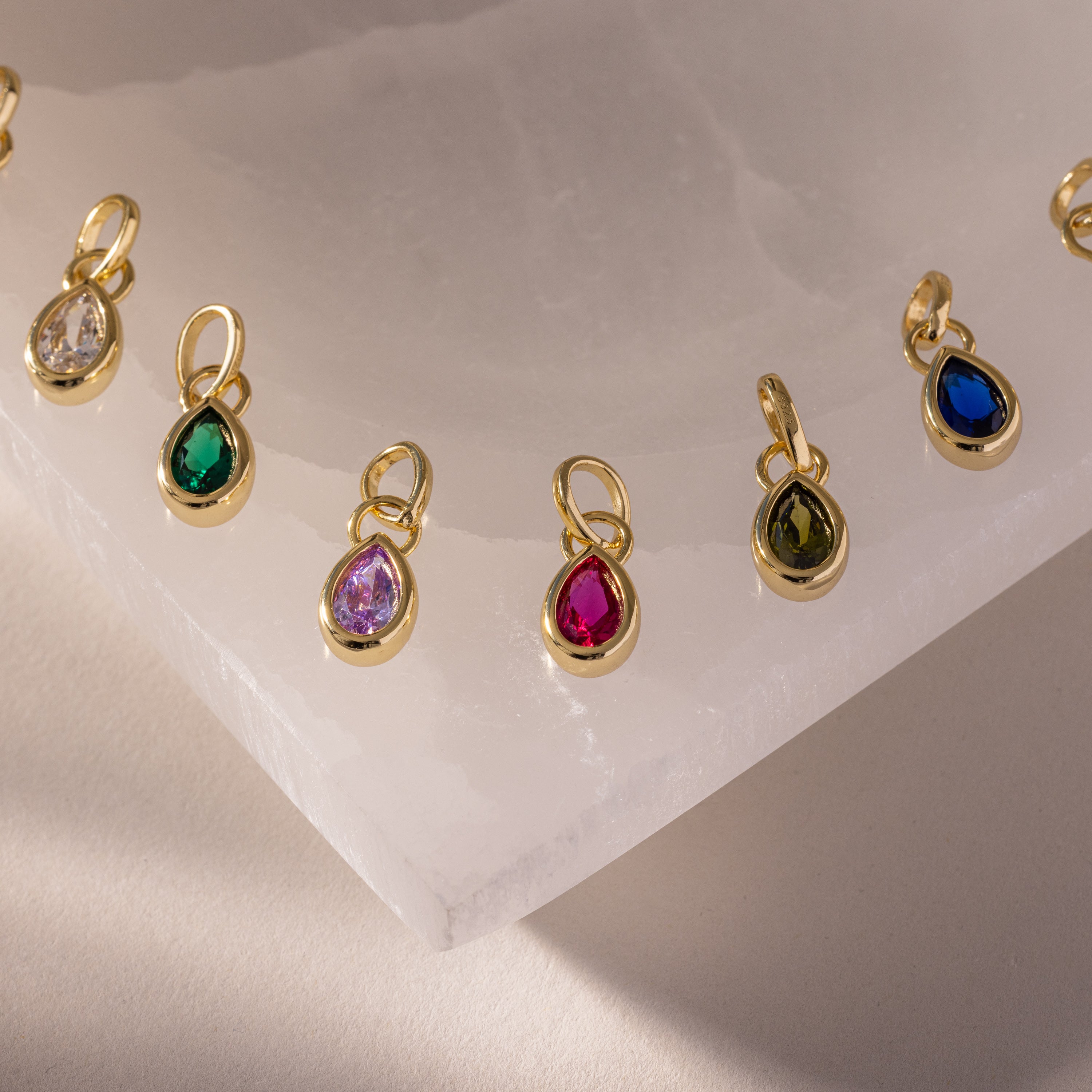 Teardrop Birthstone Charm pendants in gold bezel settings, elegantly displayed on white marble.