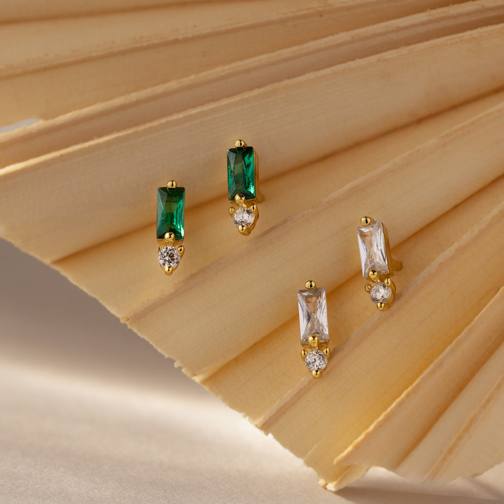 The Baguette Topaz Drop Studs showcase rectangular green and clear stones with a round diamond accent, creating a modern earring design elegantly presented on a beige fan.
