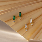 The Baguette Topaz Drop Studs showcase rectangular green and clear stones with a round diamond accent, creating a modern earring design elegantly presented on a beige fan.