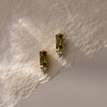 Baguette Peridot Drop Studs feature rectangular green peridot stones and small round white gems, elegantly displayed on textured off-white paper.