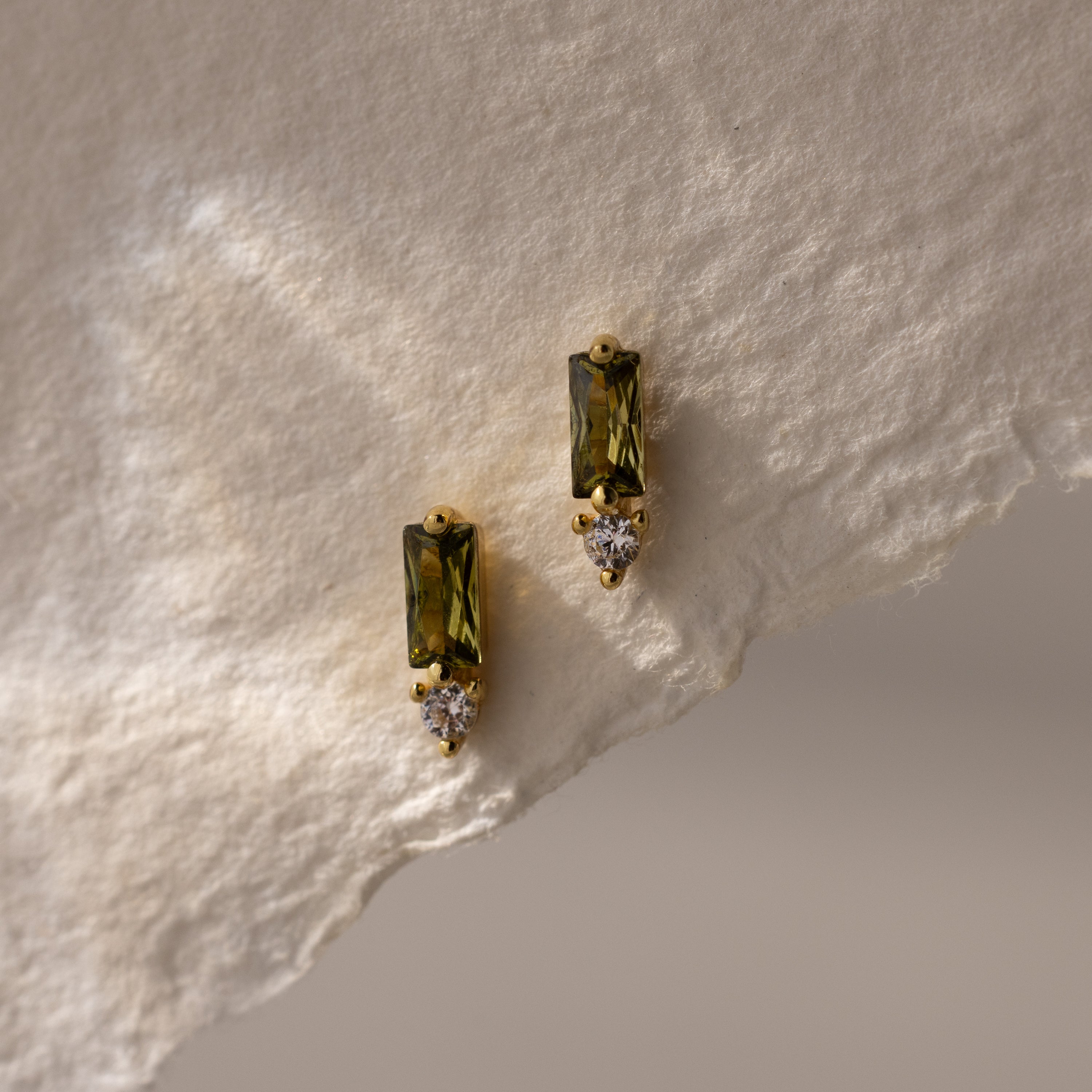 Baguette Peridot Drop Studs feature rectangular green peridot stones and small round white gems, elegantly displayed on textured off-white paper.