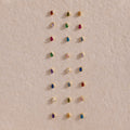 Teardrop Sapphire Flatback Studs are displayed in neat rows on a textured beige surface, showcasing a charming array of colorful birthstone jewelry.