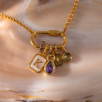 The Teardrop Birthstone Charm gold chain necklace features four charms—a K, amethyst teardrop birthstone, star, and round gem in a gold bezel—against a shell background. A perfect addition to your gemstone jewelry collection.