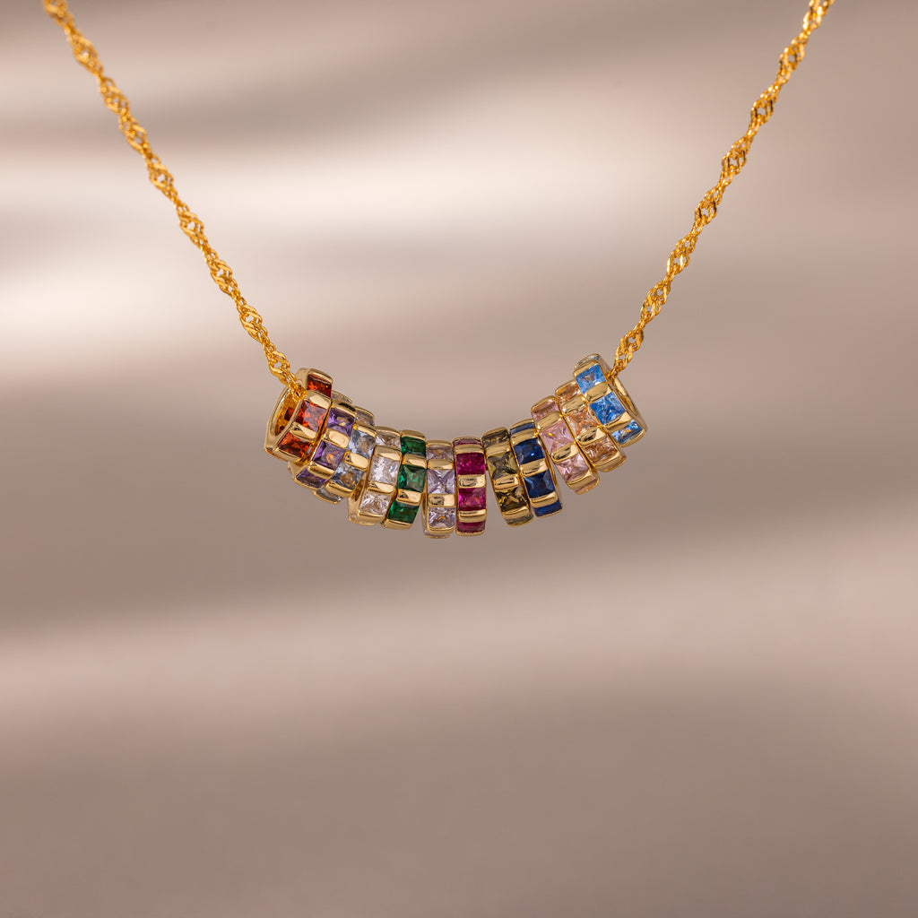 The Infinity Princess Pink Tourmaline Charm features a gold chain necklace with colorful gemstone rings, including a sparkling princess-cut tourmaline, arranged at the center and displayed on a neutral background.