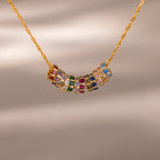 The Infinity Princess Pink Tourmaline Charm features a gold chain necklace with colorful gemstone rings, including a sparkling princess-cut tourmaline, arranged at the center and displayed on a neutral background.