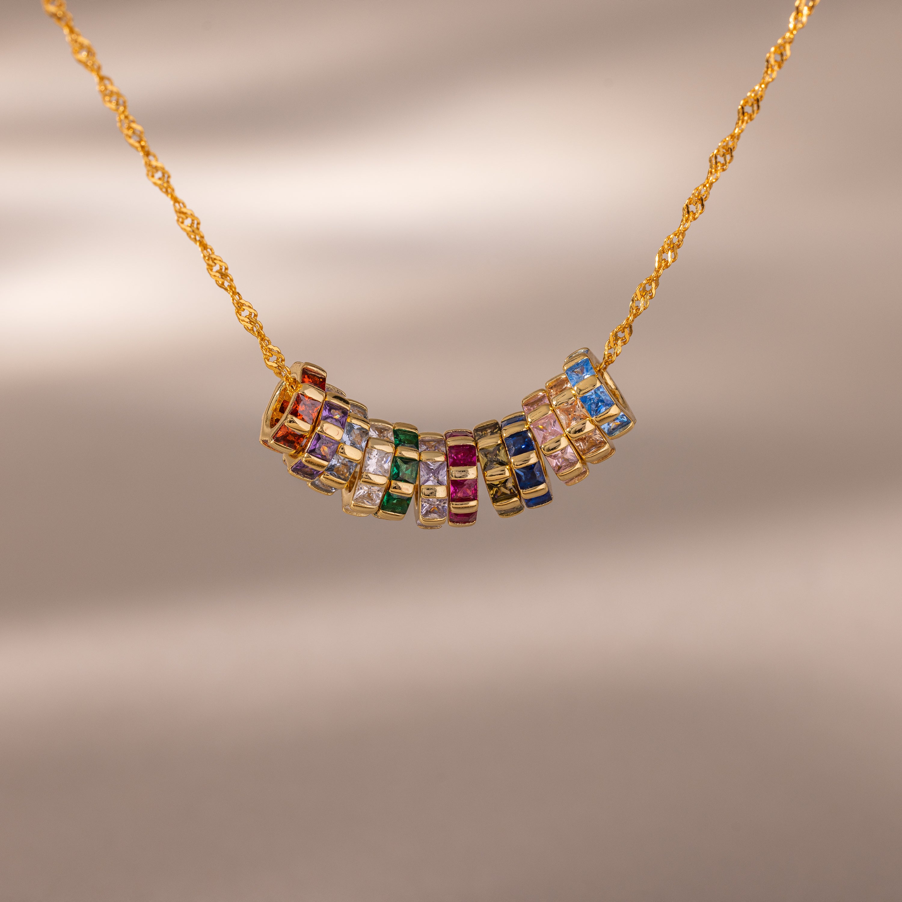 The Infinity Princess Pink Tourmaline Charm features a gold chain necklace with colorful gemstone rings, including a sparkling princess-cut tourmaline, arranged at the center and displayed on a neutral background.