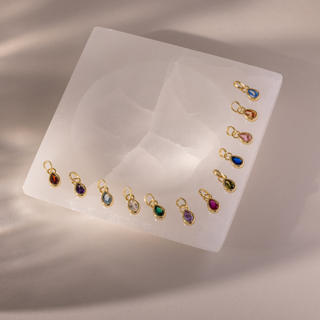 Teardrop Birthstone Charms are arranged on a square white stone slab, illuminated by soft overhead lighting that creates gentle shadows.