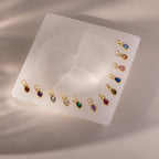 Teardrop Birthstone Charms are arranged on a square white stone slab, illuminated by soft overhead lighting that creates gentle shadows.