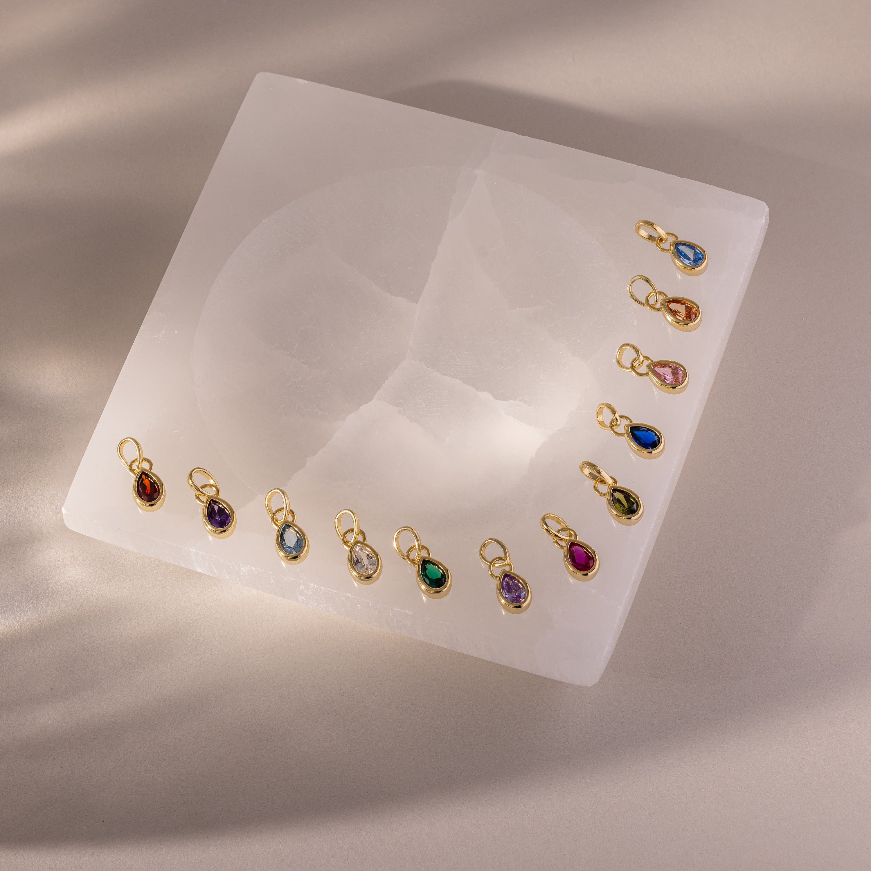Teardrop Birthstone Charms are arranged on a square white stone slab, illuminated by soft overhead lighting that creates gentle shadows.