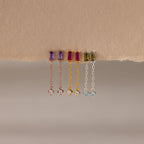 Four pairs of Duo Birthstone Drop Studs, each with vibrant baguette-cut gemstones and chain studs, hang elegantly from a torn piece of beige paper.