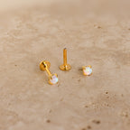 Two Opal Flat Back Studs in 18K Gold, adorned with iridescent opals, rest on a beige stone surface—ideal for anyone seeking dainty earrings with subtle elegance.
