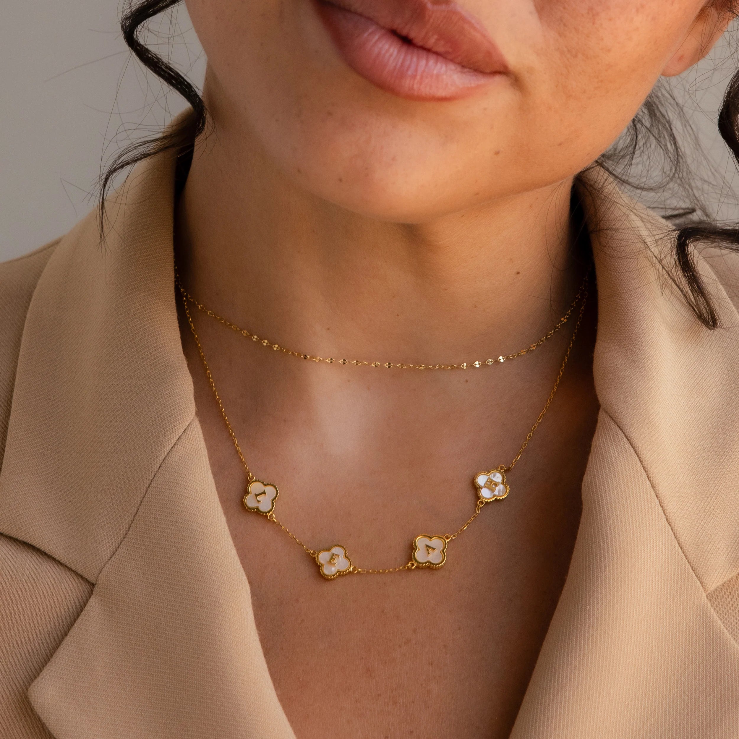 A woman in a beige blazer models the Gemstone Clover Letter Necklace, featuring four flower-shaped pendants, along with a sleek gold chain.