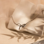 The Herringbone Bow Necklace in Sterling Silver rests on a wavy, cream ceramic dish—perfect for adding a touch of coquette style to any look.