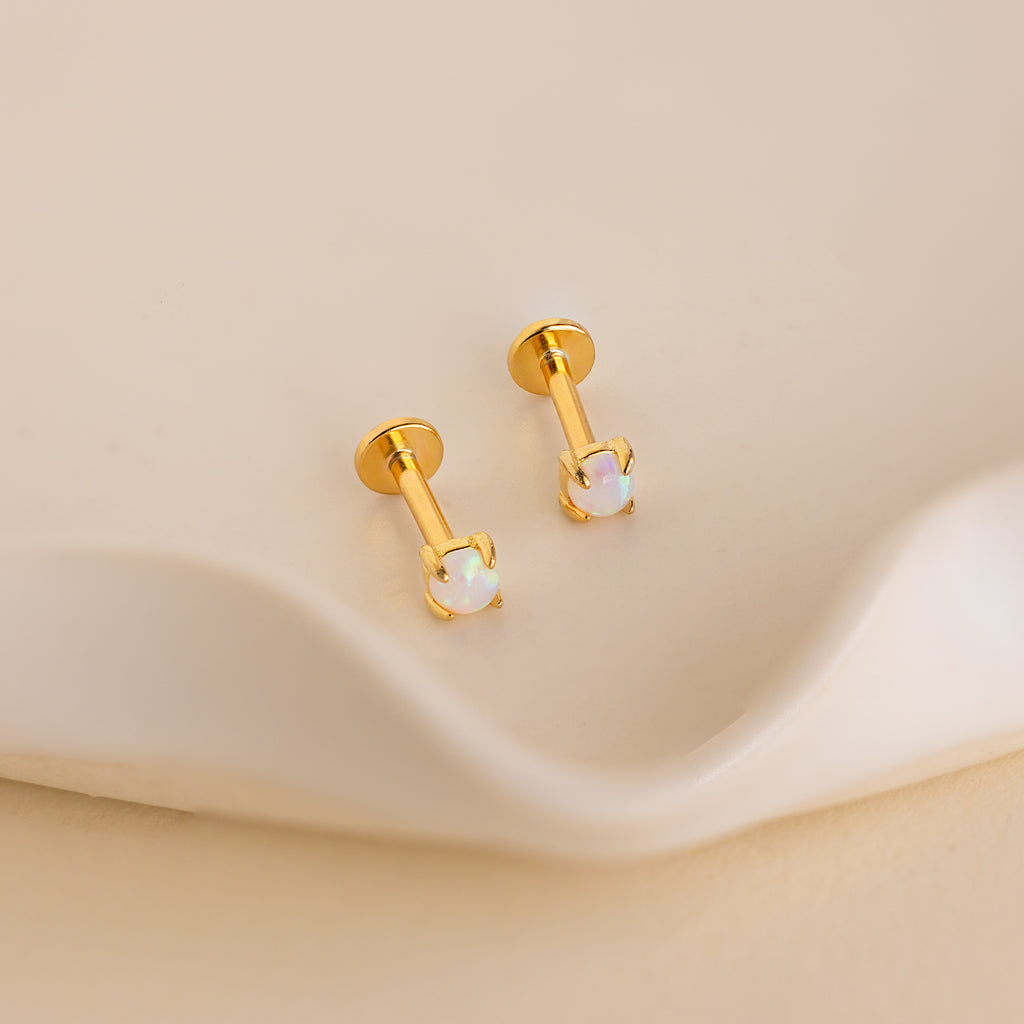 A pair of Opal Flat Back Studs in 18K Gold sits on a smooth, cream surface—these dainty earrings offer a subtle, elegant touch to any style.