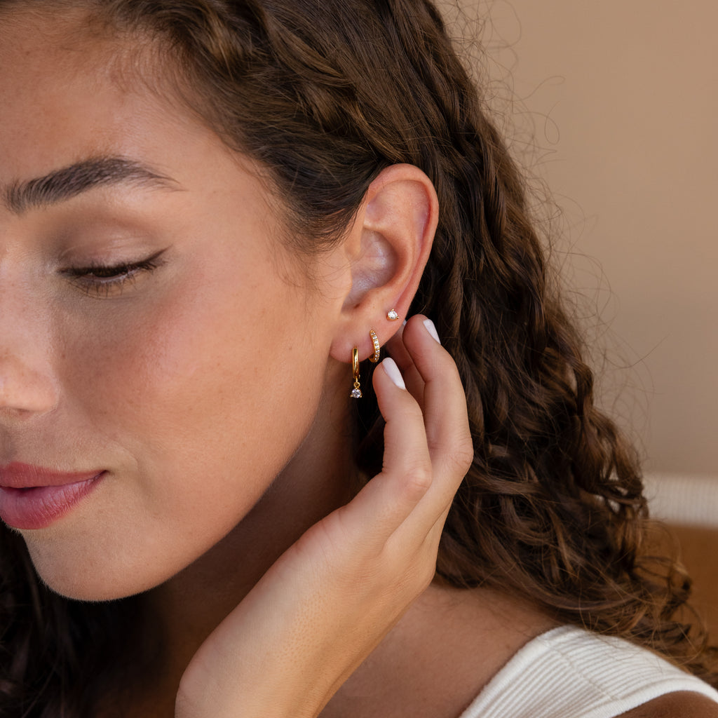 A woman with curly hair touches her ear, showcasing the Classic Diamond Earrings Set—multiple gold earrings including a classic stud—while wearing a white top.