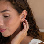 A woman with curly hair touches her ear, showcasing the Classic Diamond Earrings Set—multiple gold earrings including a classic stud—while wearing a white top.