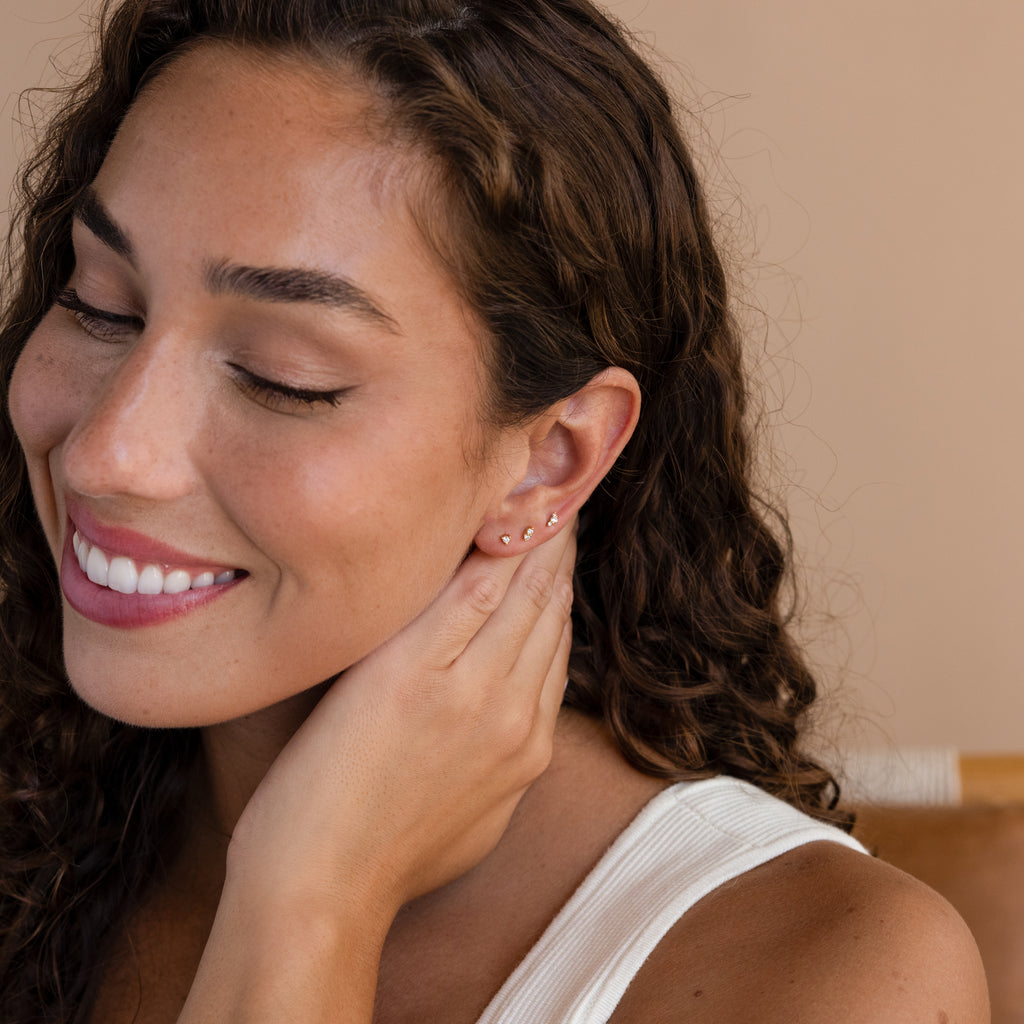 A woman with her eyes closed, wearing Tiny Diamond Flatback Studs set in 18K Gold, exudes quiet elegance.