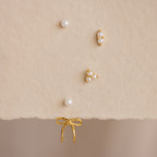 The Pearl & Bow Ear Jackets in 18K Gold, featuring five assorted gold and pearl studs including elegant bow designs, are displayed on a soft beige textured surface.