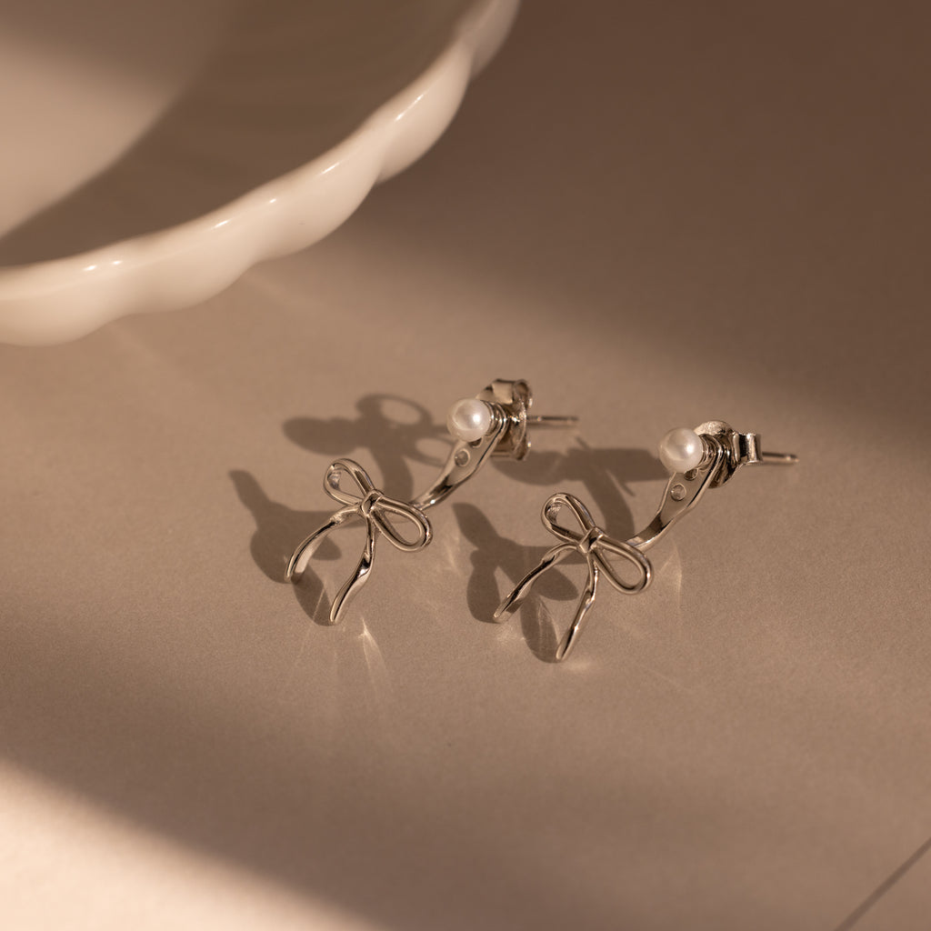 The Pearl & Bow Ear Jackets in Sterling Silver feature delicate pearl accents in a unique bow design, resting elegantly on a neutral surface near a white ceramic dish.