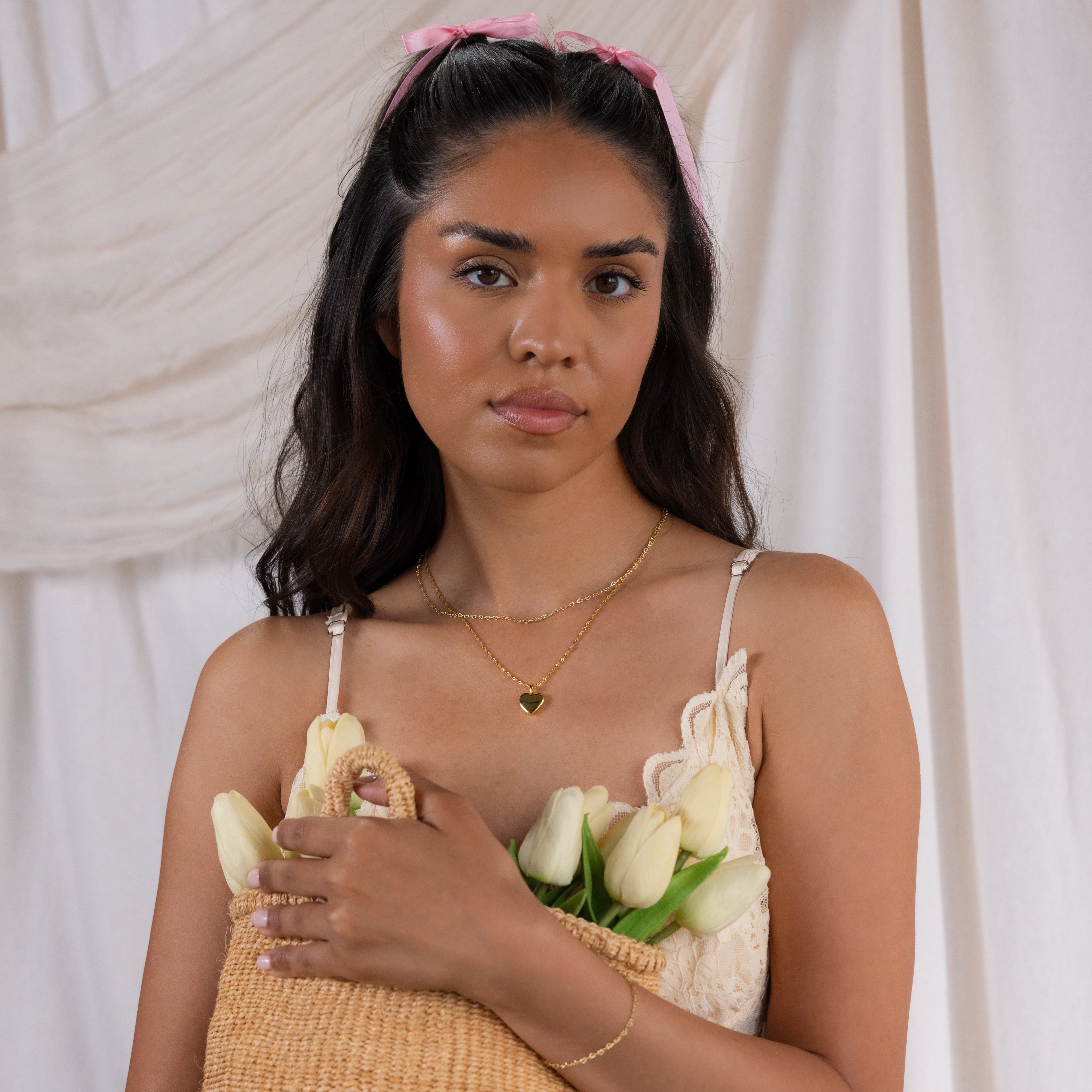 Woman with long dark hair and a pink bow holds a straw bag filled with white tulips, wearing the Heart Link Chain Necklace, against a soft fabric backdrop.