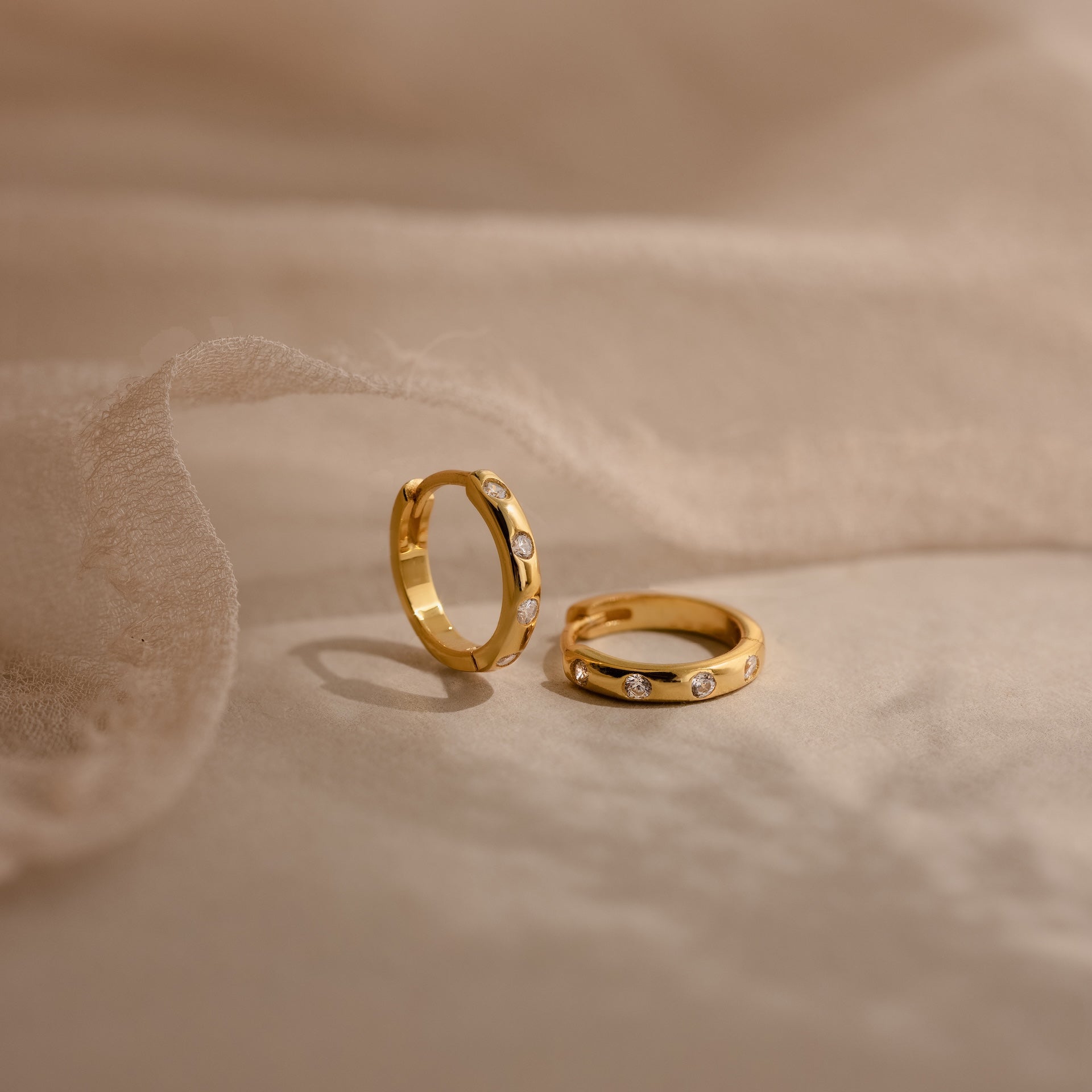 Two gold rings with small diamonds rest on soft beige fabric, styled alongside Mini Diamond Sprinkle Huggies in 18K Gold for a timeless, elegant look.