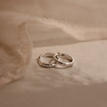 Two Mini Diamond Sprinkle Huggies in Silver rest on a soft beige fabric background, their sparkling diamonds evoking the elegance of delicate silver hoops.