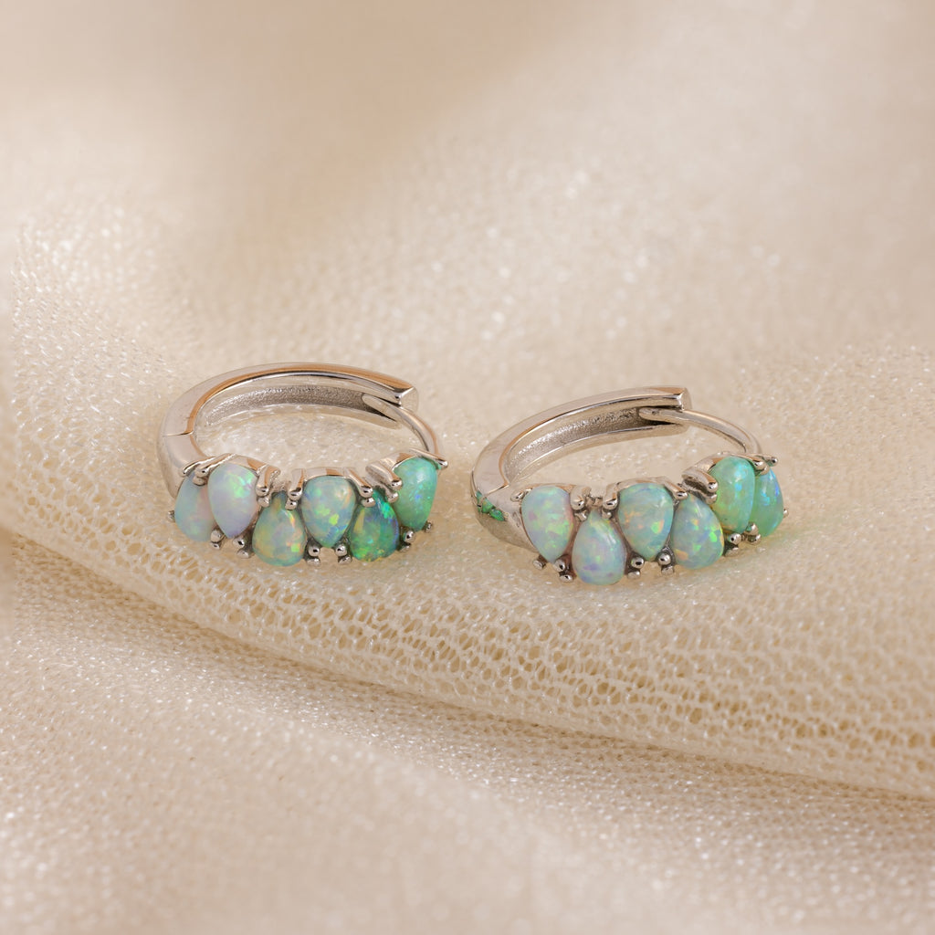 A pair of Ombre Opal Huggies in Silver with small oval opal gems, elegantly displayed on a beige fabric background.