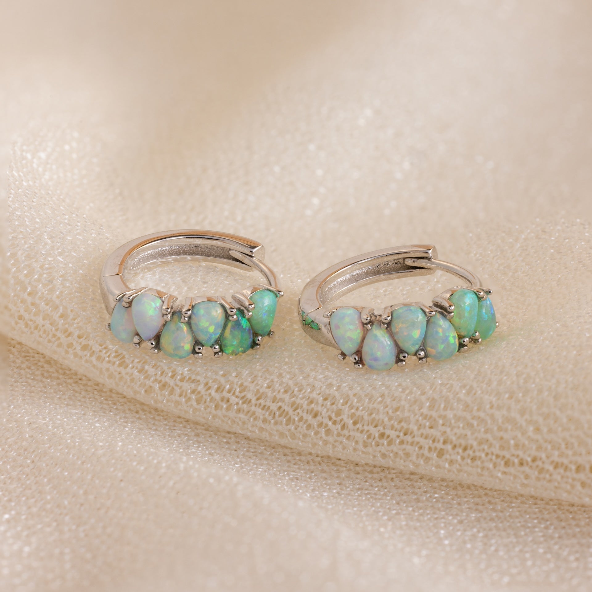 A pair of Ombre Opal Huggies in Silver with small oval opal gems, elegantly displayed on a beige fabric background.