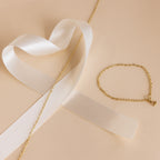 Gold chain bracelet with a heart charm beside a white satin ribbon on beige, perfect for pairing with our Heart Link Chain Necklace.