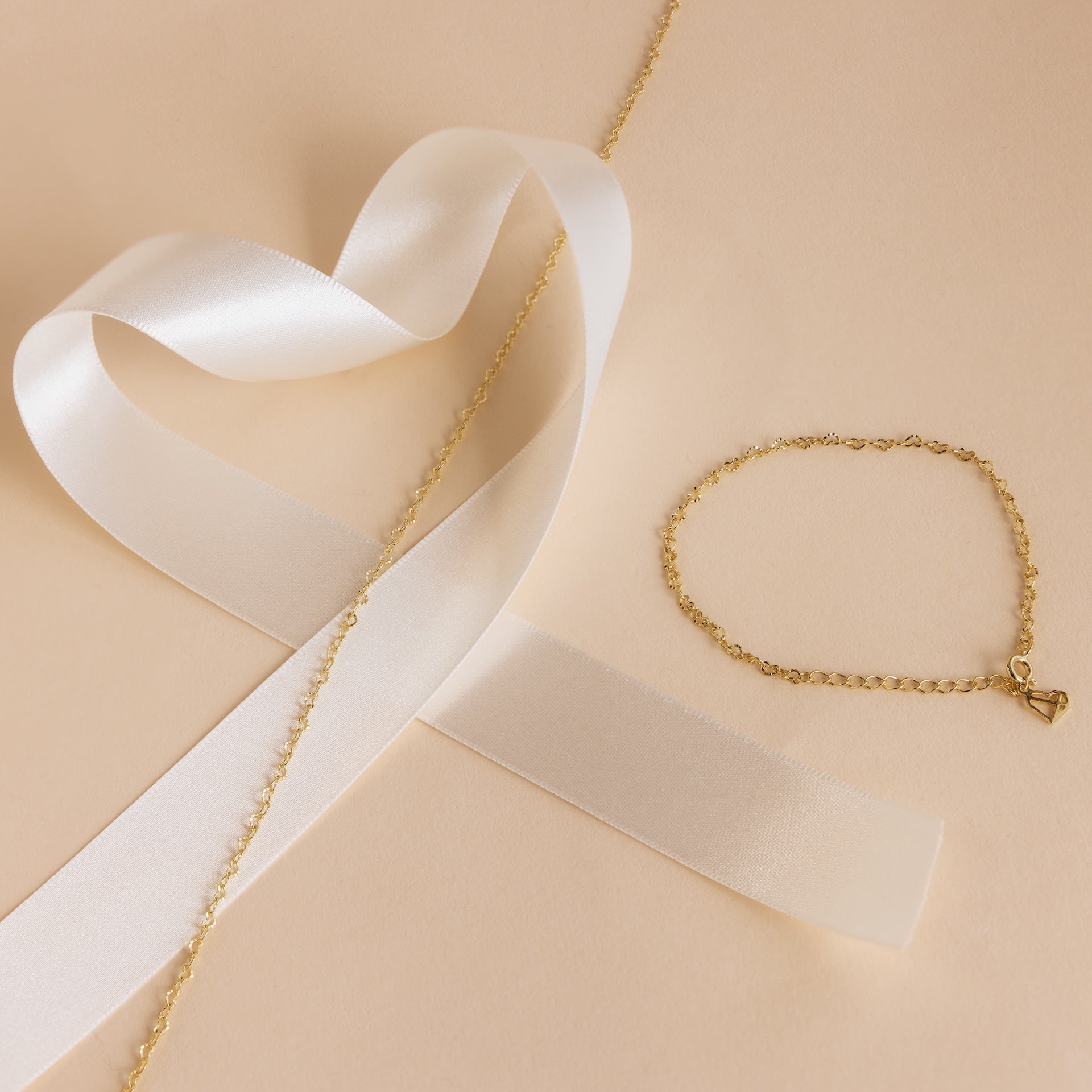 Gold chain bracelet with a heart charm beside a white satin ribbon on beige, perfect for pairing with our Heart Link Chain Necklace.