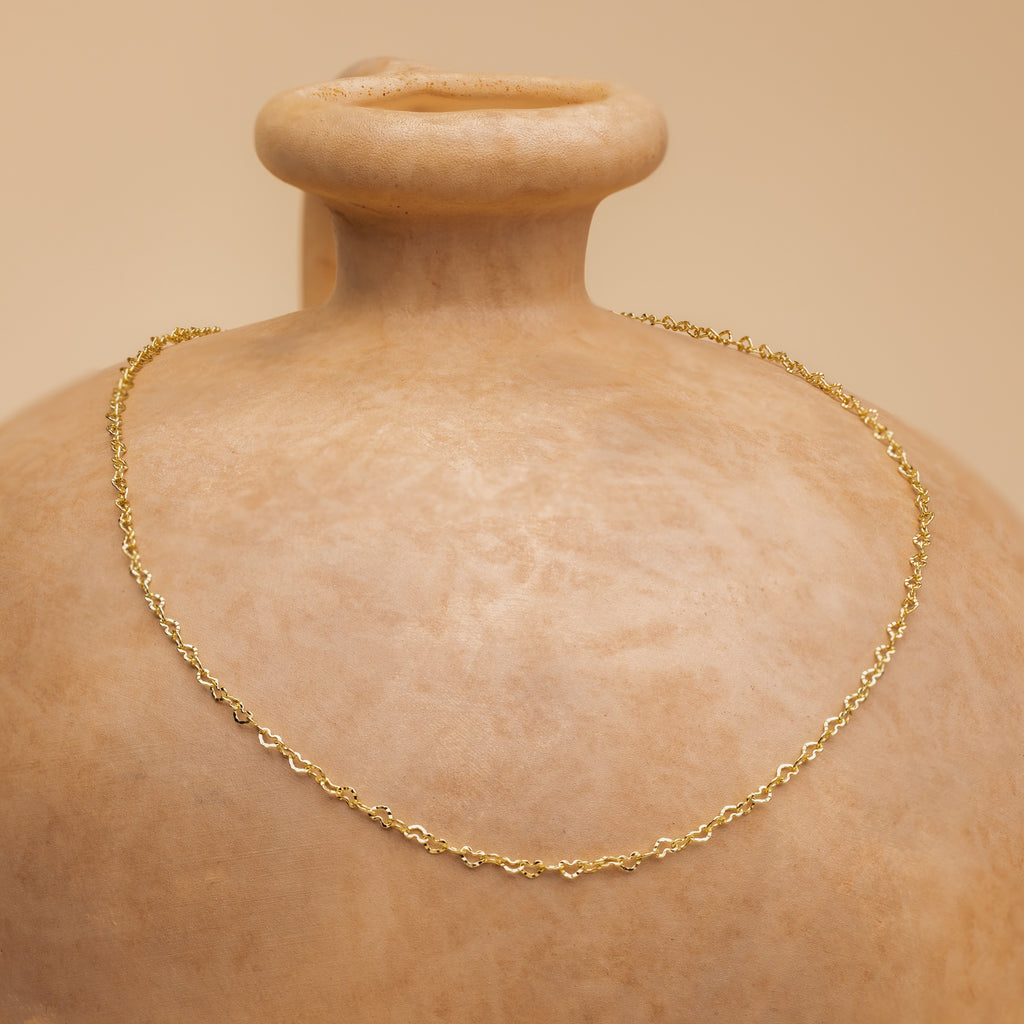 The Heart Link Chain Necklace, with its interlocking hearts, is displayed on a smooth clay vase against a beige background, adding a subtle touch of romance.