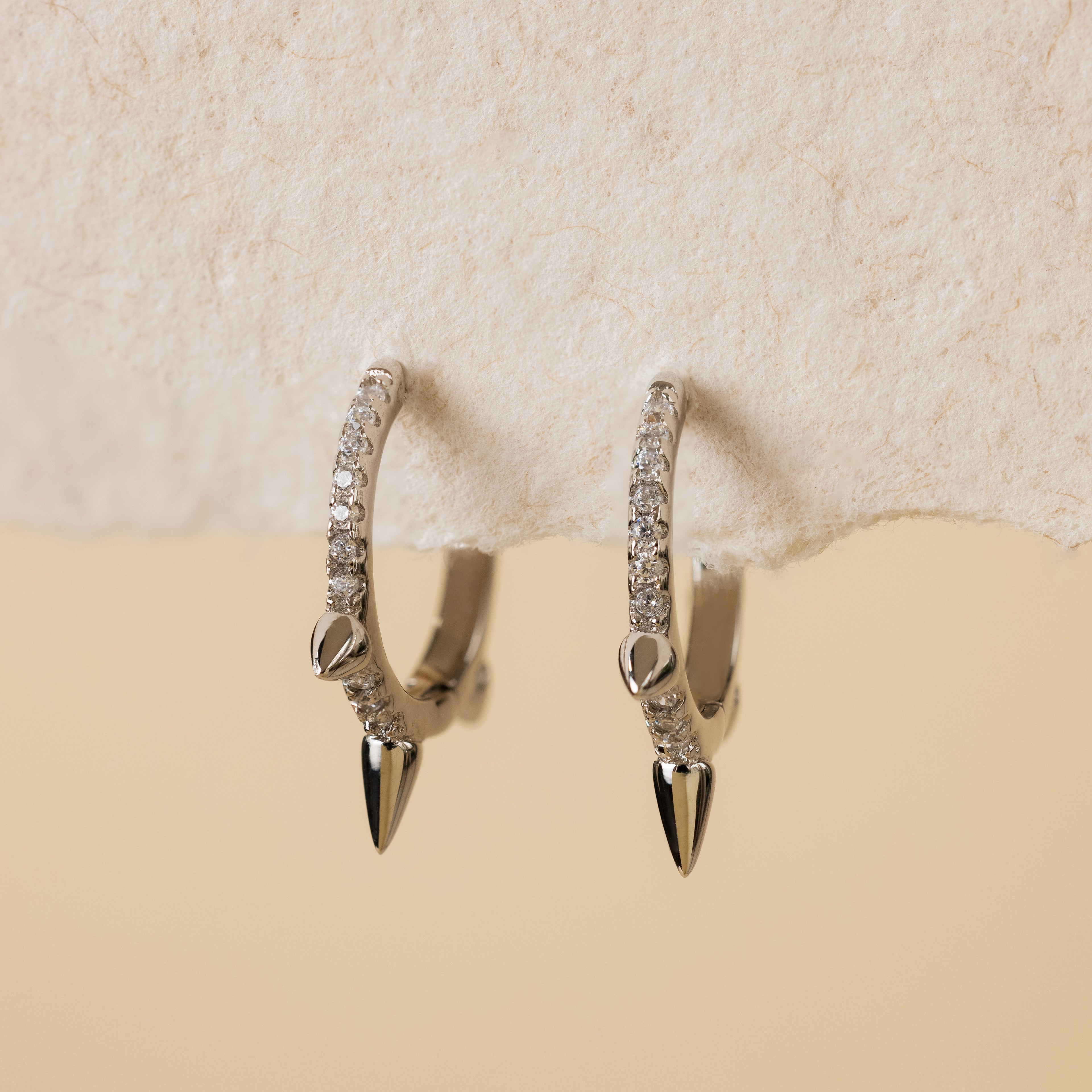 Minimalist jewelry: Pave Triple Spike Huggies—silver hoop earrings with small clear stones and pointed tips, shown on textured off-white paper.