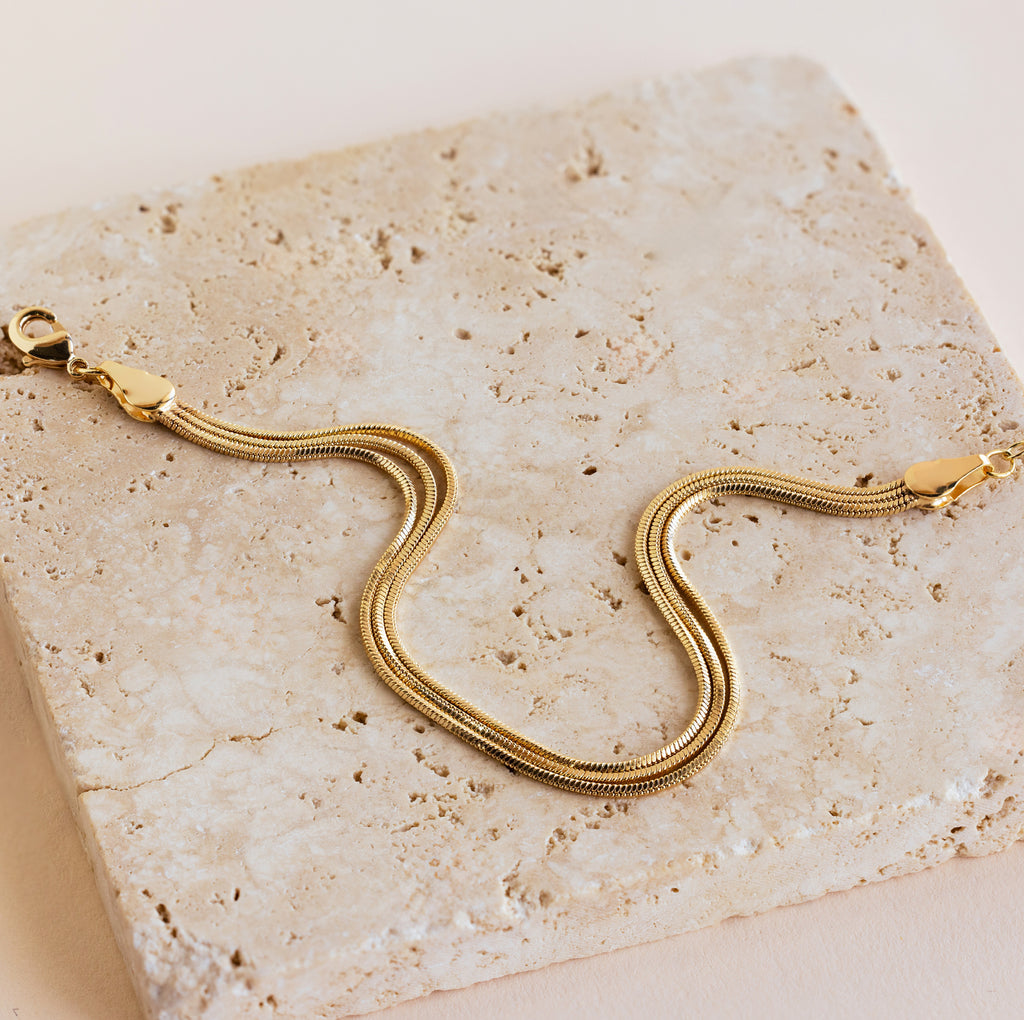 The Triple Snake Chain Bracelet features a lobster clasp and is displayed on a textured beige stone—ideal for minimal jewelry lovers or layering for a bold look.