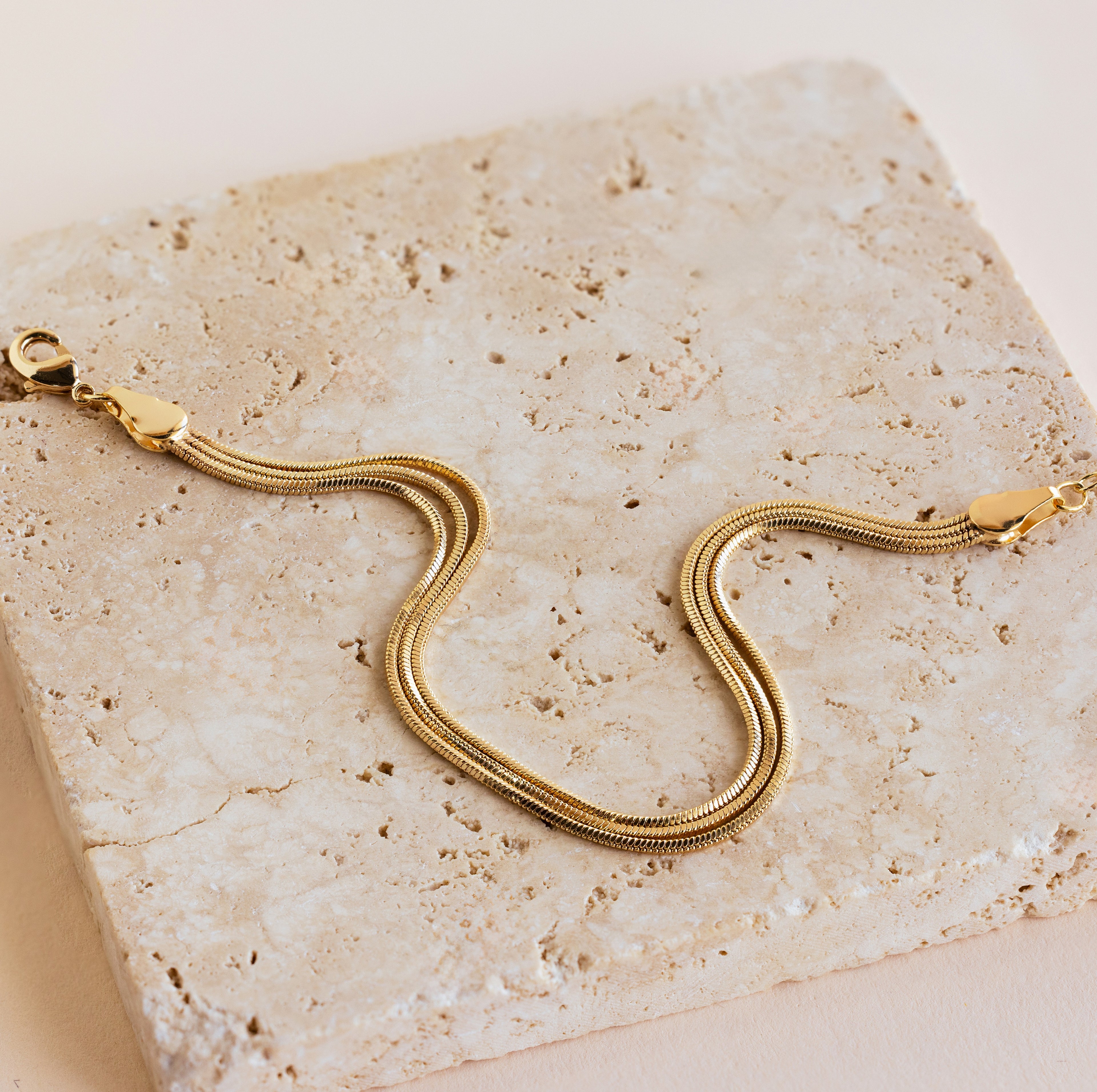 The Triple Snake Chain Bracelet features a lobster clasp and is displayed on a textured beige stone—ideal for minimal jewelry lovers or layering for a bold look.
