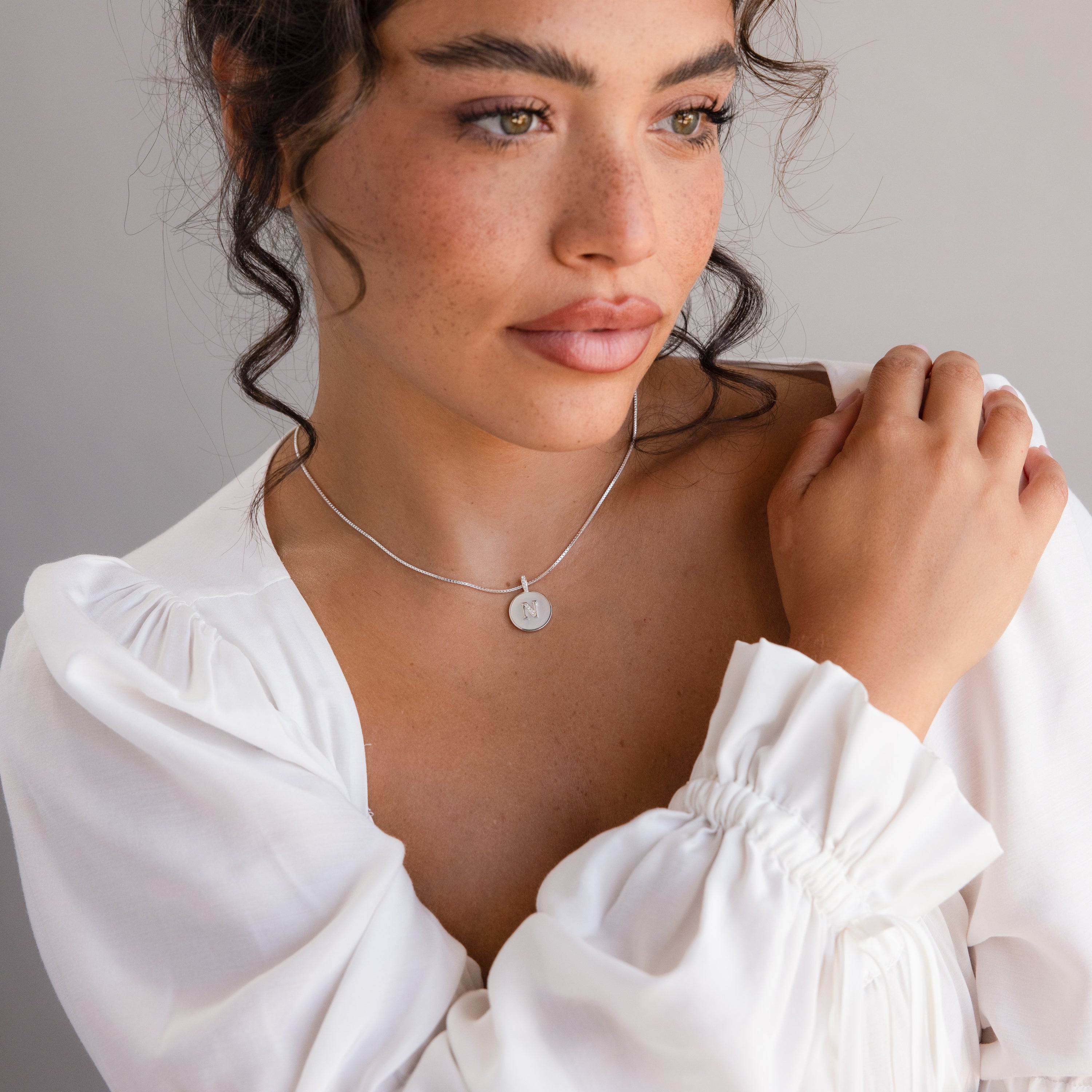 Woman with curly hair wearing a white blouse and the Pearl Initial Disc Necklace, gazing to the side.